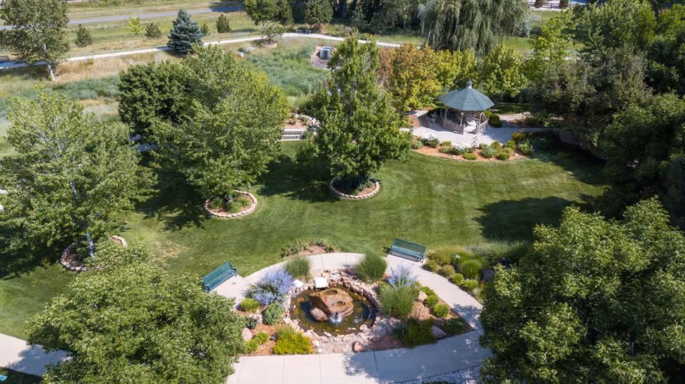 Aerial view of a landscaped garden area with green grass, several trees, a small pond with rocks, two green benches, and a gazebo surrounded by shrubs and flowers.