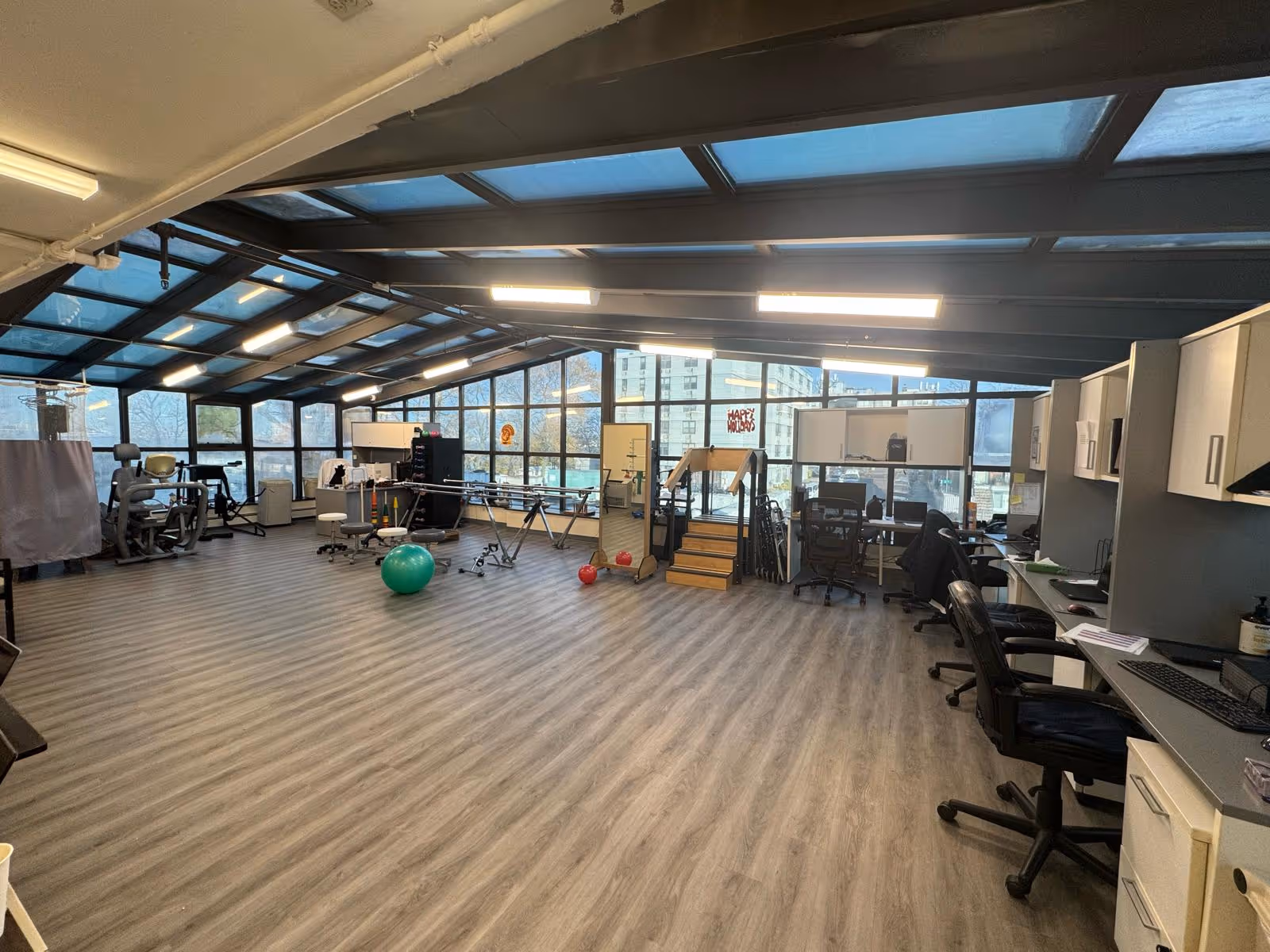 A spacious rehabilitation or therapy room with large windows and a glass ceiling allowing natural light to fill the space. The room has exercise equipment including a green exercise ball, parallel bars, and a set of stairs for physical therapy. There are office desks and chairs along one side with computers and cabinets. The floor is covered with wood-patterned vinyl or laminate.