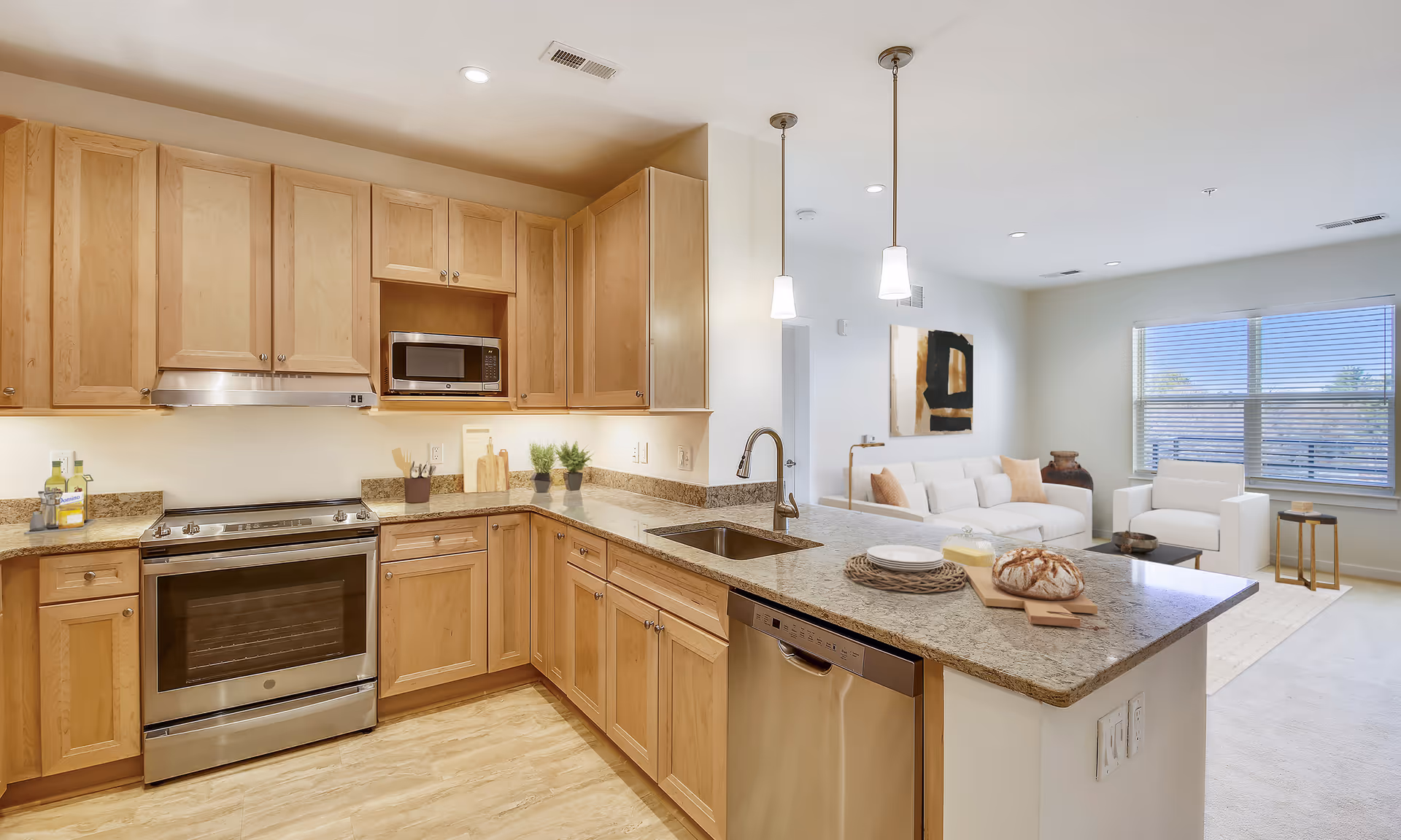 Open-concept kitchen with wooden cabinets, a granite island and sink overlooking a bright living area with a sofa and large window.