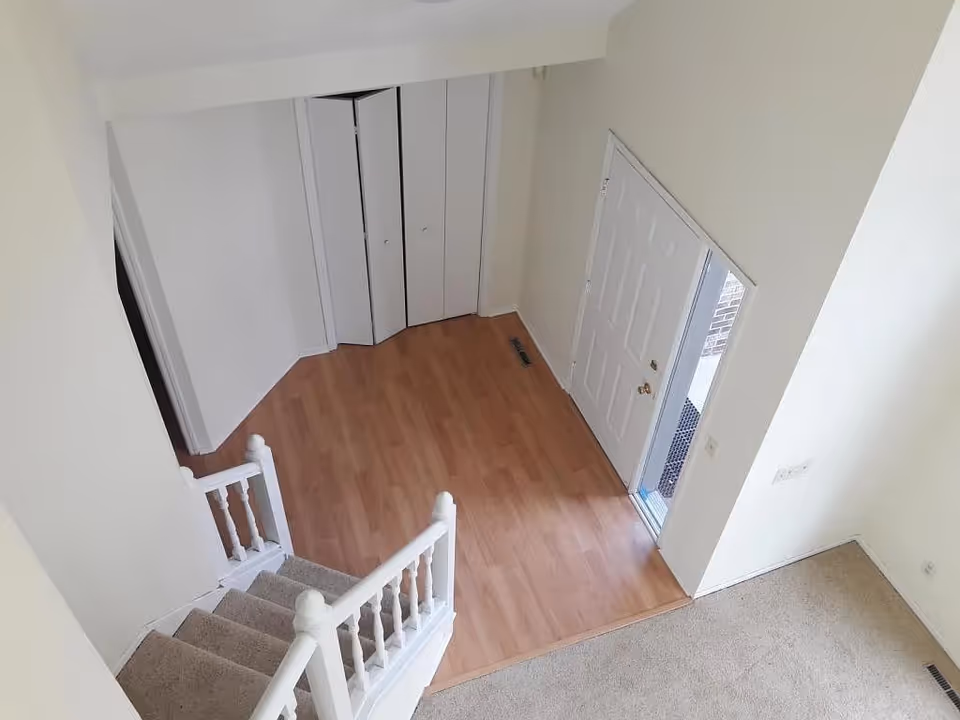 View from the top of a carpeted staircase looking down into an entryway with light wood flooring, a white front door, and a closet with white bi-fold doors. The walls are painted light beige and there is a carpeted area adjacent to the entryway.