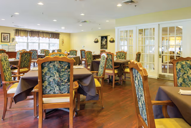 A dining room with multiple wooden tables covered with dark tablecloths and surrounded by wooden chairs with floral patterned upholstery. The room has large windows with curtains, light yellow walls, and a wooden floor. There are decorative wall hangings and a set of glass-paneled double doors.