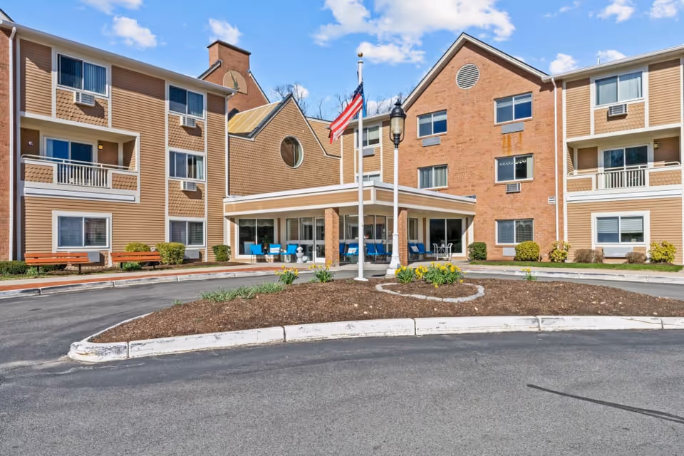 Exterior view of a senior living facility building with a circular driveway, landscaped center island with flowers, and an American flag on a flagpole. The building has multiple windows, balconies, and a covered entrance with seating.
