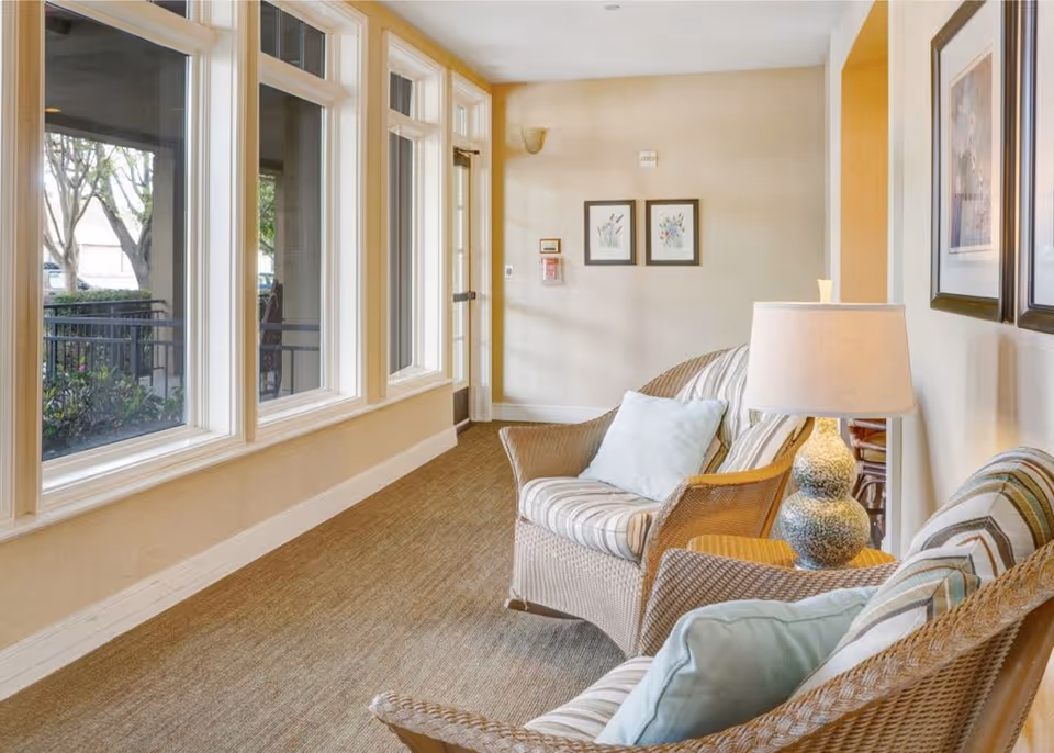 A cozy indoor seating area with wicker chairs featuring striped cushions and light blue pillows, a side table with a decorative lamp, large windows letting in natural light, and framed artwork on the walls.