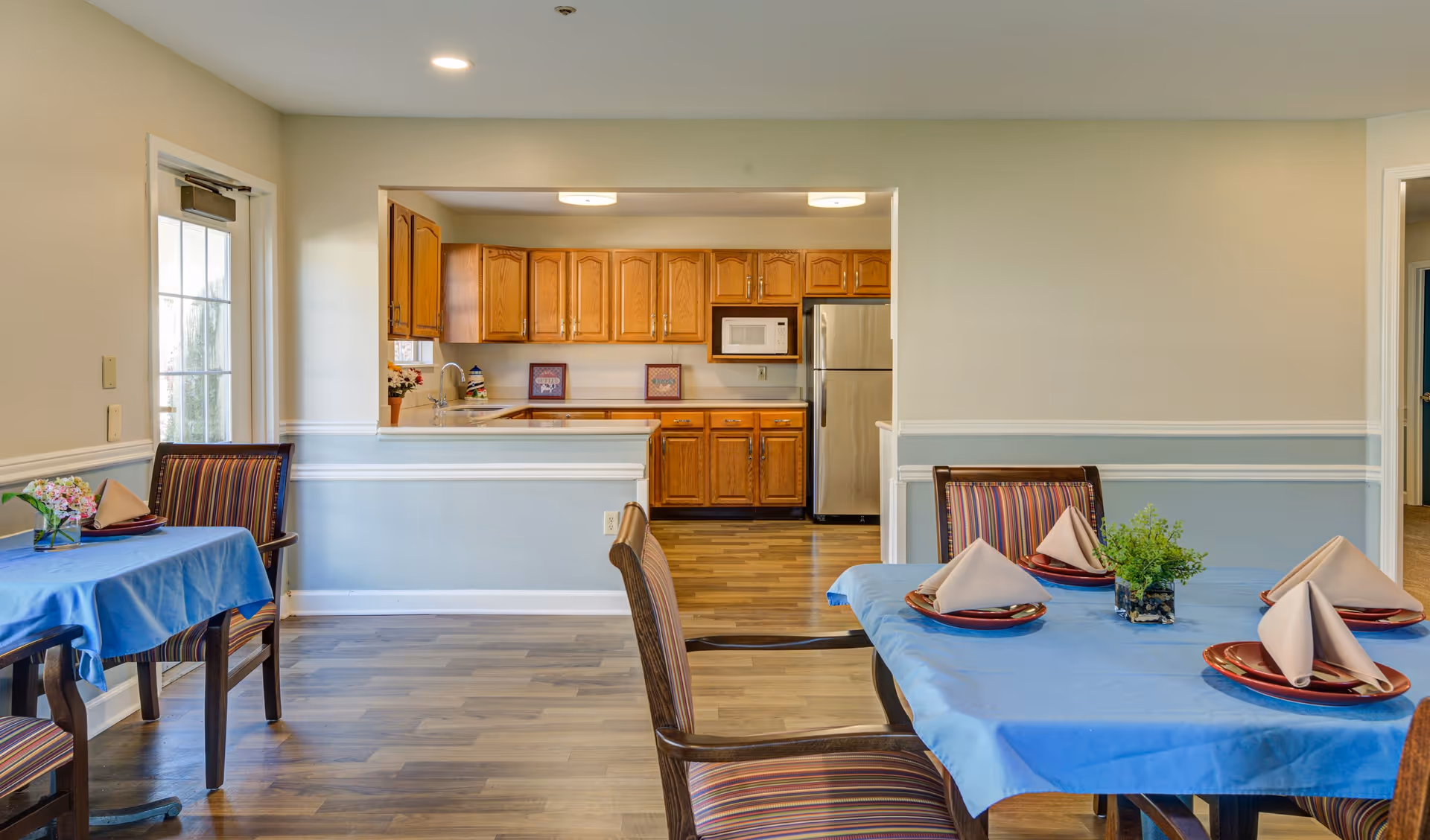 A dining area with two tables covered in blue tablecloths, each set with red plates and folded beige napkins. There are striped cushioned chairs around the tables. In the background, there is a kitchen with wooden cabinets, a microwave, and a stainless steel refrigerator. The walls are painted beige with a light blue wainscoting, and the floor is wood.