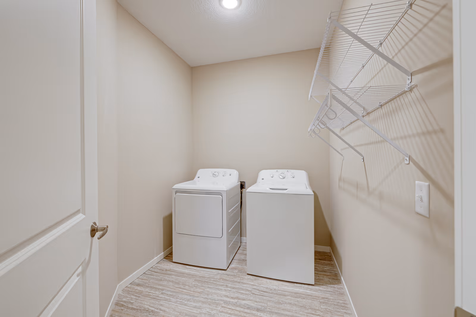 A small laundry room with a white washing machine and dryer side by side against a beige wall. Above the machines is a white wire shelf for storage. The room has light-colored flooring and a closed white door on the left.