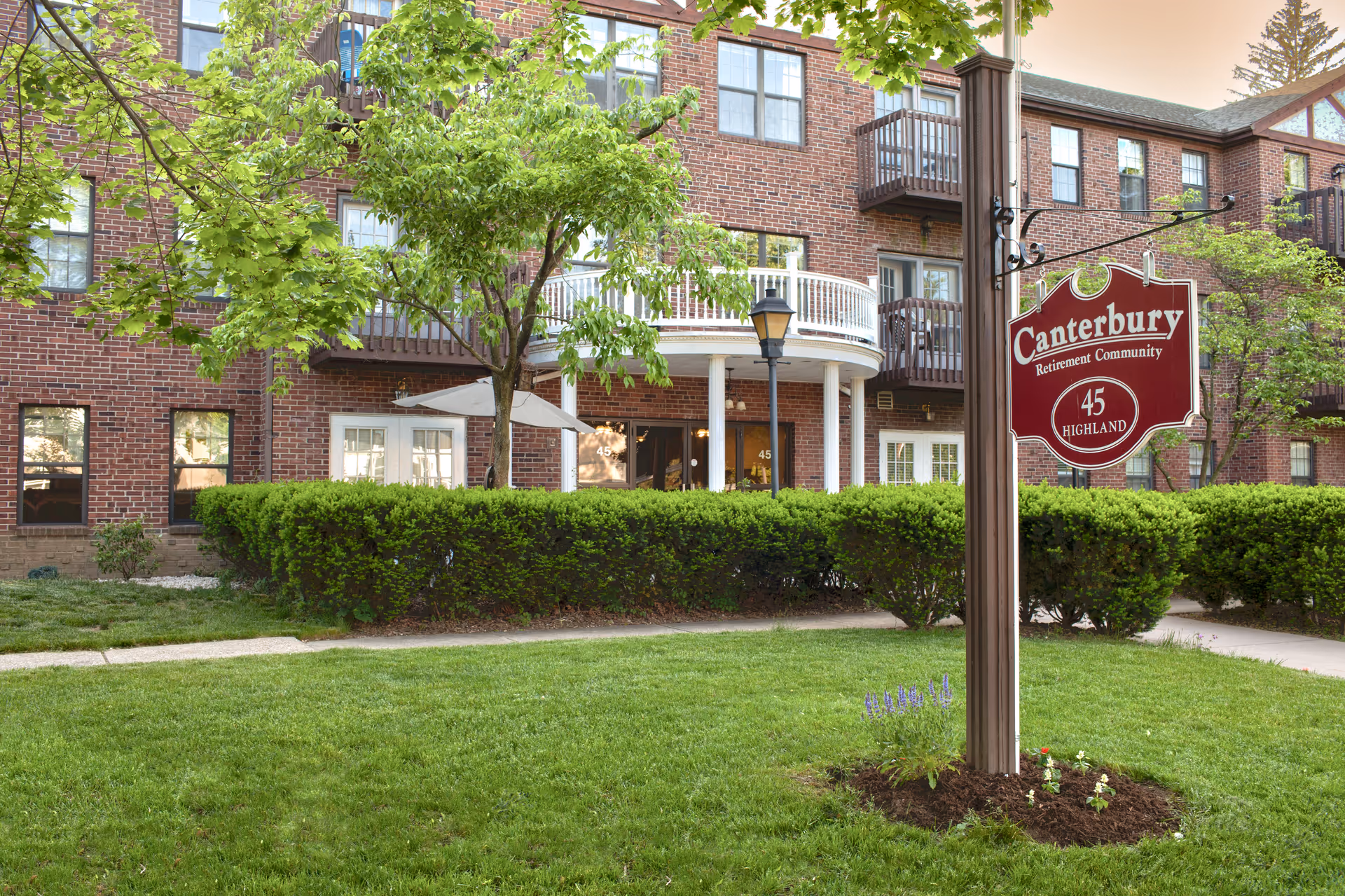 Exterior view of Canterbury Retirement Community building with red brick walls, multiple windows, balconies, green bushes, trees, and a lawn. A red sign with white text reads 'Canterbury Retirement Community 45 Highland' mounted on a post in the foreground.