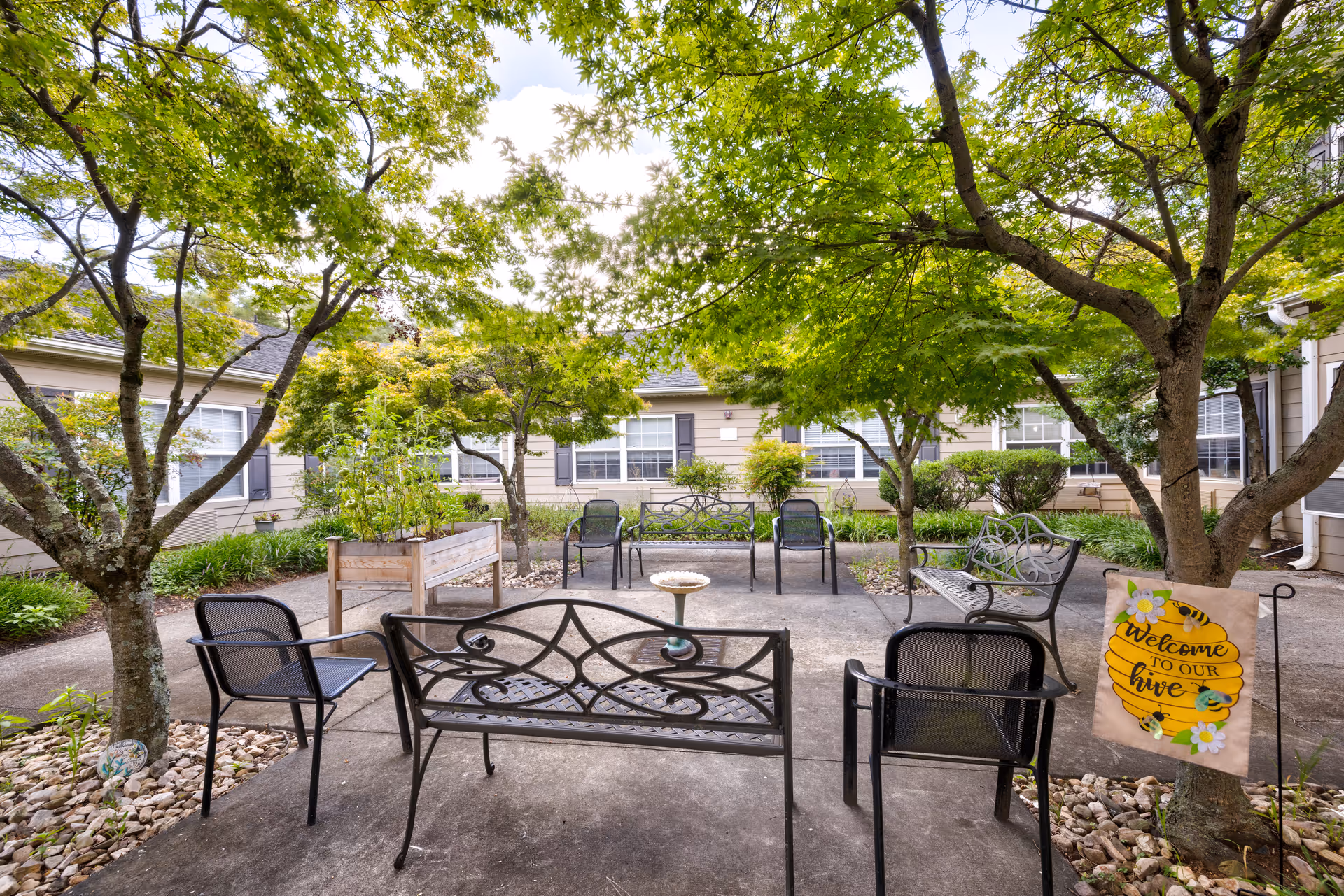 Outdoor courtyard area with several black metal benches and chairs arranged around a small birdbath. The space is shaded by green leafy trees and surrounded by a beige building with multiple windows. A small garden bed with plants and rocks is visible, along with a decorative sign hanging from a tree that reads 'Welcome to our hive' with bee and flower illustrations.