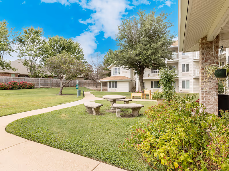Courtyard with concrete picnic tables and winding walkways in front of a multi-story senior living building under a blue sky.