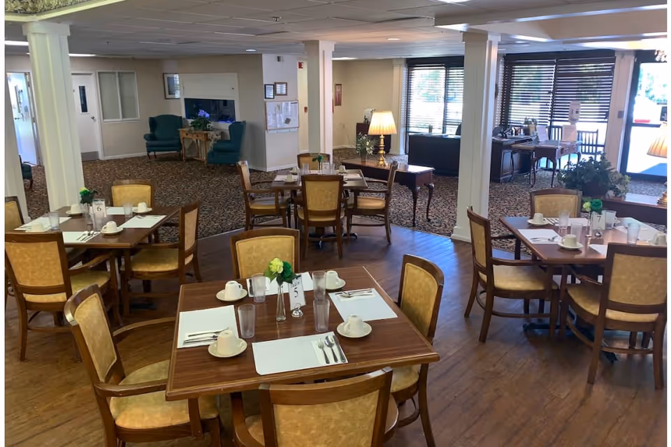 Dining area in Oakview Terrace Assisted Living and Memory Care with several wooden tables and chairs set with placemats, cups, glasses, and silverware. The room has wood flooring and carpeted sections, with large windows letting in natural light. There are lamps and some greenery decorating the space.