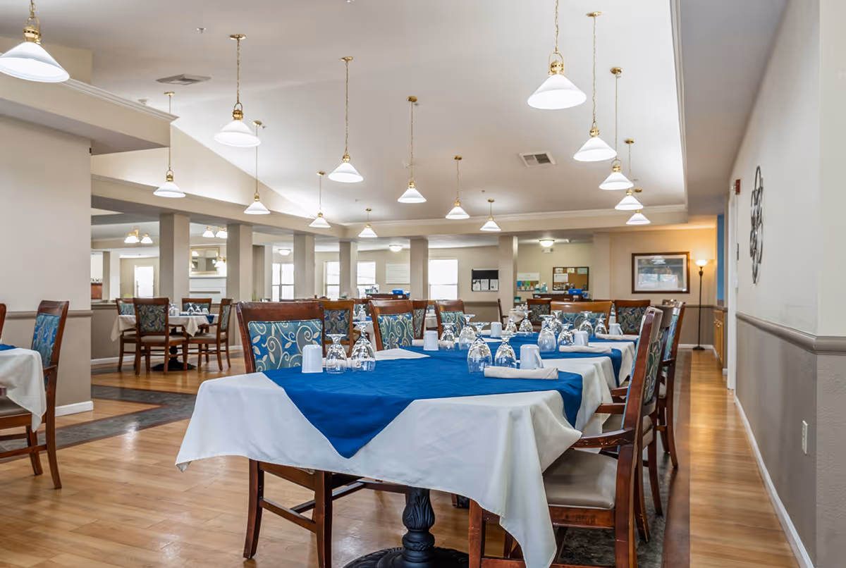 A spacious dining room with multiple tables covered in white tablecloths and blue table runners. Each table is set with upside-down glasses and napkins. The room features wooden chairs with patterned blue cushions, hardwood floors, and multiple hanging pendant lights from a high ceiling. The walls are light-colored with some framed artwork and a floor lamp in the background.
