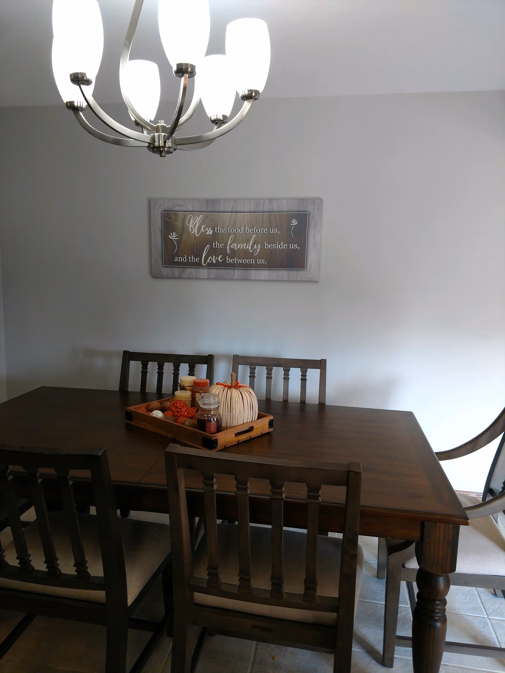 A dining room with a dark wooden table and six chairs. On the table is a wooden tray holding decorative items including candles and a pumpkin. Above the table hangs a modern chandelier with six white glass shades. A framed sign on the wall reads, 'Bless the food before us, the family beside us, and the love between us.'