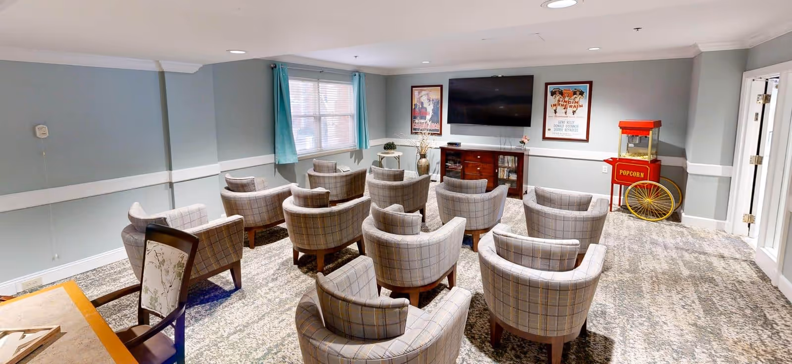 A small theater-style room with multiple plaid upholstered chairs arranged in rows facing a large flat-screen TV mounted on the wall. The room has light blue walls with white trim, a window with blue curtains, a popcorn machine in the corner, and framed movie posters on the wall.