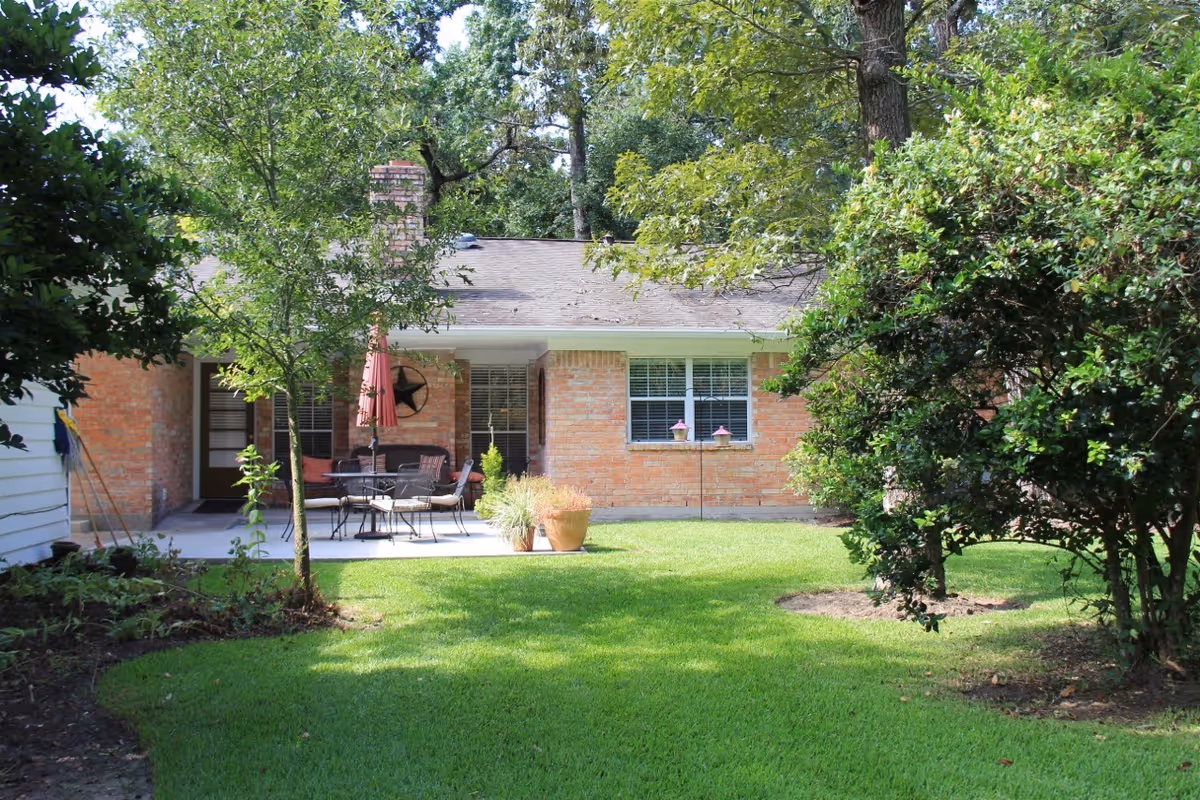 Brick single-story cottage exterior with a backyard patio, green lawn, trees, and outdoor seating with an umbrella.