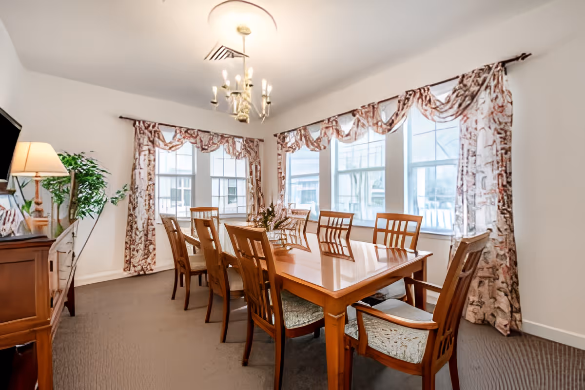 Dining room with a long wooden table and several chairs under a chandelier beside large curtained windows.