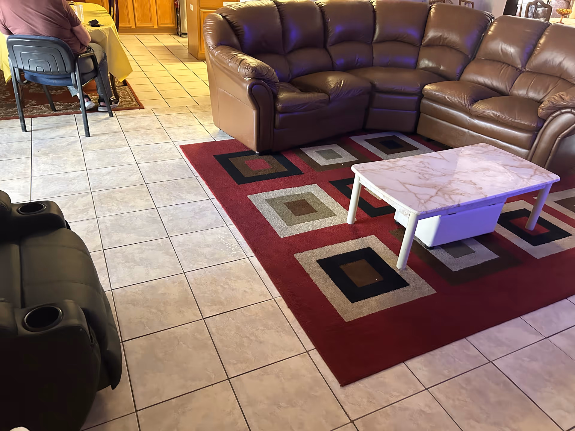 Interior view of a living room area with a brown leather sectional sofa on a red patterned rug, a white marble-top coffee table, and a black recliner chair. In the background, a person is seated at a table covered with a yellow tablecloth near a kitchen area with wooden cabinets.