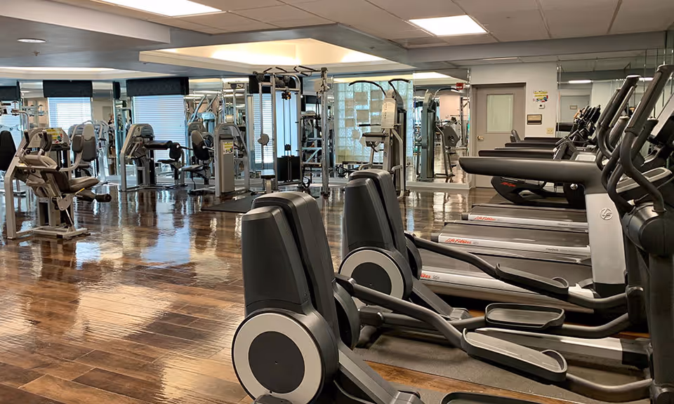 Empty fitness center with treadmills, ellipticals and weight machines on a glossy wood floor and mirrored walls.