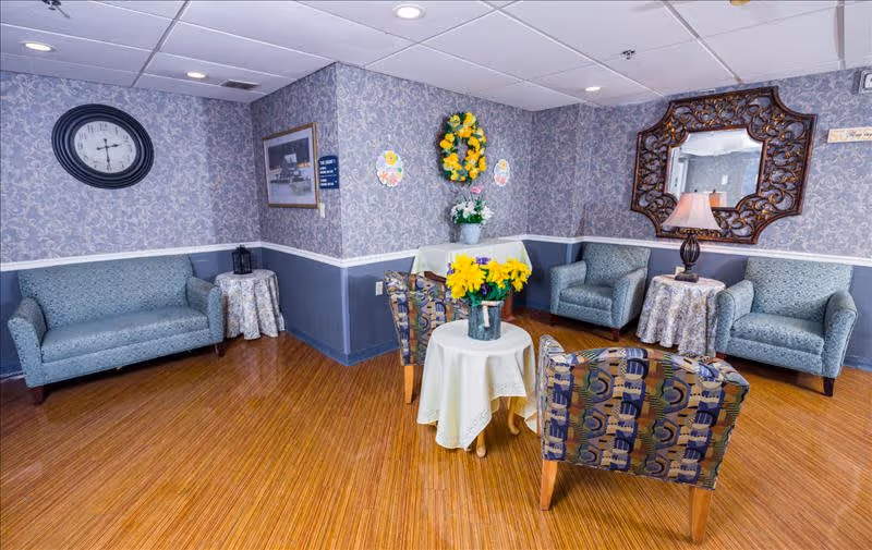 A cozy sitting area with patterned wallpaper and wooden flooring. The room features a blue sofa, two blue armchairs, and two patterned chairs arranged around small tables covered with white cloths. A large decorative mirror and a table lamp are on one side, while a clock and framed picture hang on the walls. A vase with yellow flowers is on the central table, and a floral wreath decorates the corner wall.
