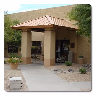 Covered outdoor seating area with two benches under a metal roof supported by four columns, surrounded by desert landscaping with gravel, potted plants, and trees.