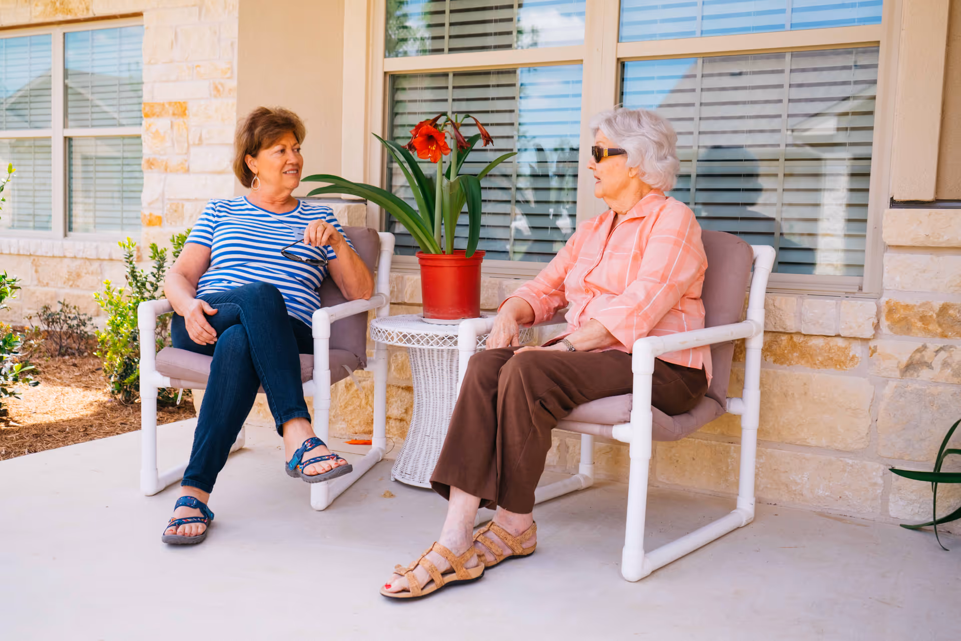Two elderly women sitting and chatting on cushioned patio chairs outside a building with stone walls and large windows. A small white wicker table with a red potted plant is between them.