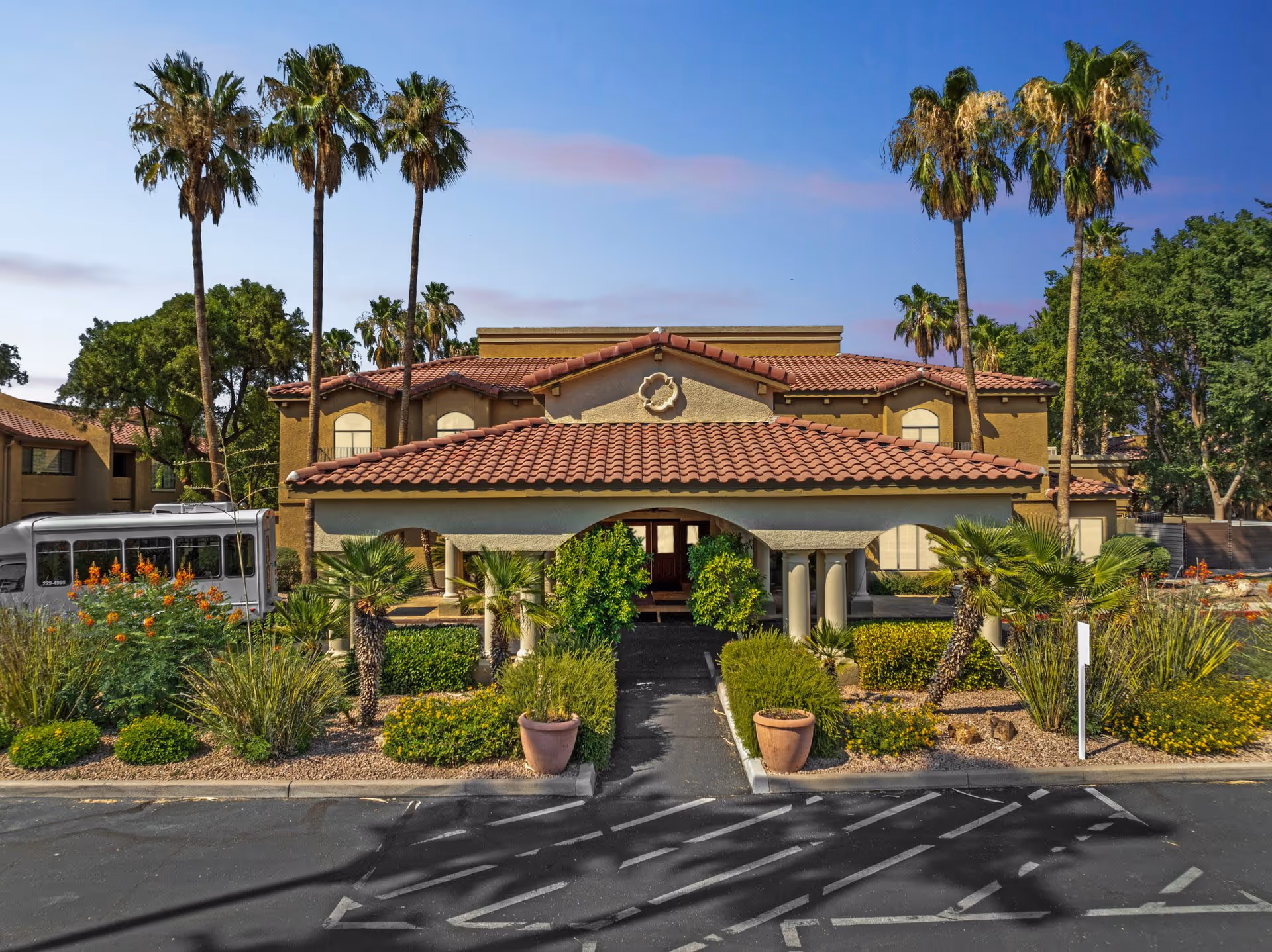 Front exterior view of a two-story building with a red tile roof, surrounded by palm trees and desert landscaping, with a covered entrance and a shuttle bus parked to the left.