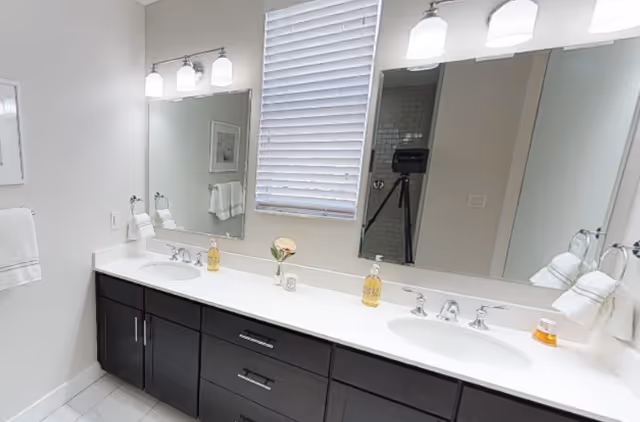 A modern bathroom vanity with a white countertop and two sinks, each with chrome faucets. Above the sinks is a large mirror with three-light fixtures mounted on the wall. There are hand towels on towel rings on both sides of the vanity, a small vase with flowers, and soap dispensers on the countertop. A window with closed blinds is centered above the vanity.