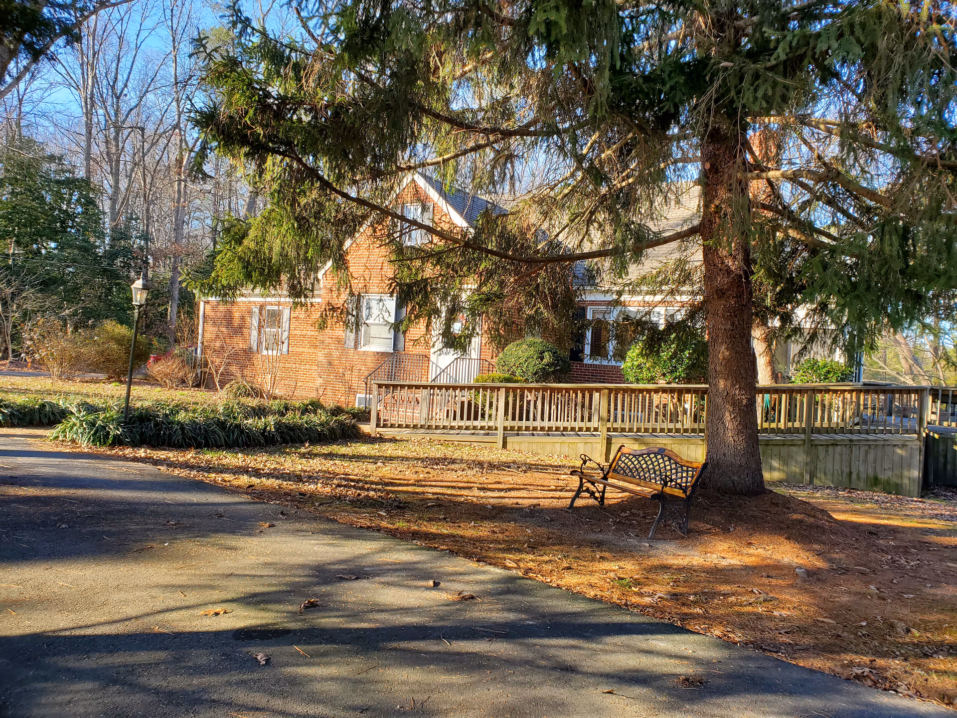 Outdoor view of a brick house partially obscured by a large evergreen tree. There is a wooden ramp leading to the house entrance and a metal bench placed under the tree. The surrounding area has some bushes, leafless trees, and a paved driveway.