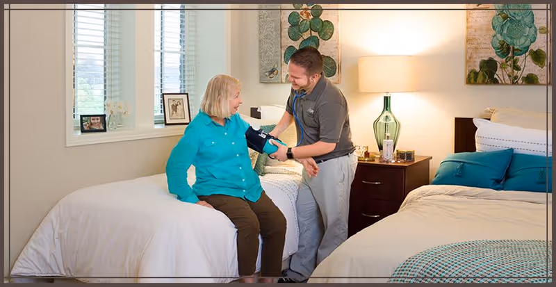 A healthcare professional is measuring the blood pressure of an elderly woman sitting on the edge of a bed in a well-lit bedroom. The room has two beds with white bedding and teal pillows, a nightstand with a lamp, and framed pictures on the wall and windowsill.