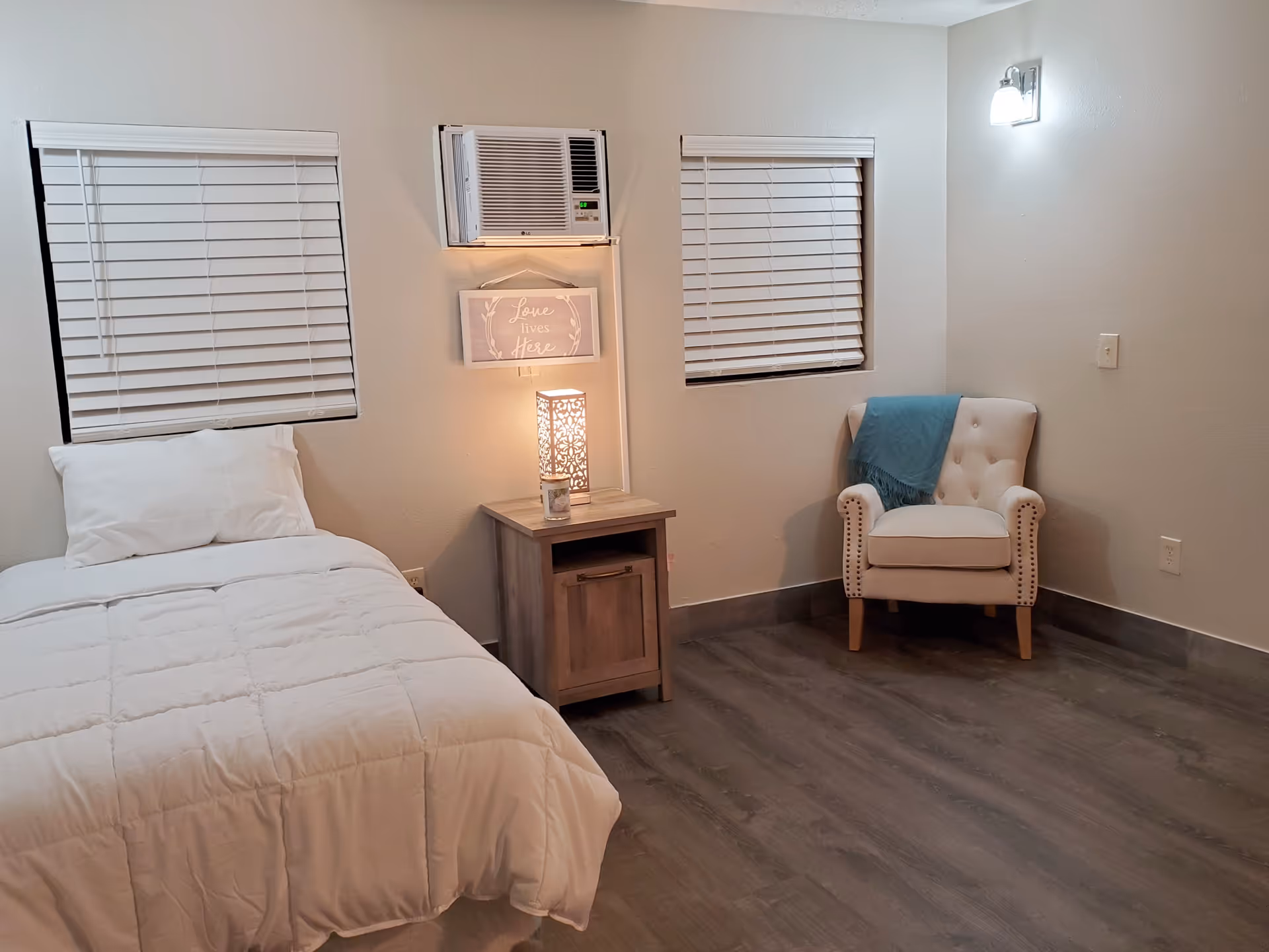A simple bedroom with a single bed covered in white bedding, a wooden nightstand with a decorative lamp and candle, two windows with white blinds, a wall-mounted air conditioner, a framed sign that reads 'Love lives Here', and a beige armchair with a blue throw blanket draped over it.