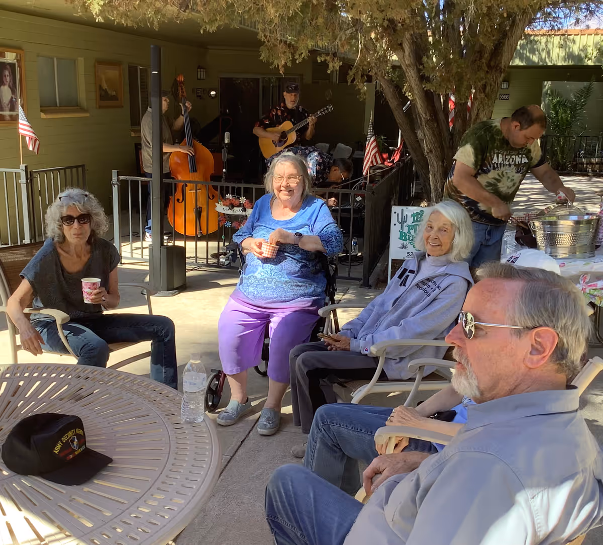 A group of older adults sitting around patio tables and enjoying drinks while a small band plays under a tree outside a senior living facility.