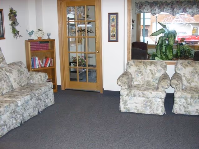 A living room area with floral patterned sofa and two matching armchairs on a carpeted floor. There is a wooden bookshelf with books and decorative items on the left side, a wooden door with glass panels in the center, and a large window with plants on the right side. A framed picture hangs on the wall between the door and window.