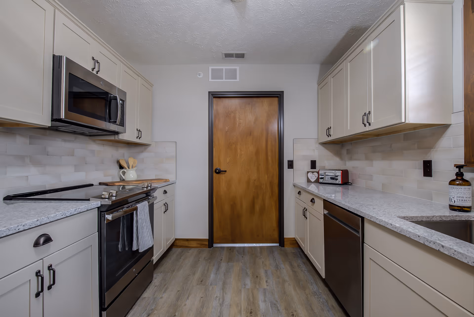 A modern kitchen with light gray cabinets, a stainless steel microwave and oven, a dishwasher, and a wooden door at the end. The countertops are light-colored with a subtle pattern, and the backsplash features rectangular tiles in neutral tones. The floor is wood-style laminate, and there are kitchen utensils and a toaster on the counters.
