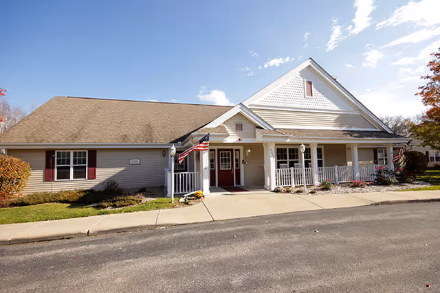 Front exterior of a single-story residential-style senior living building with a covered porch, American flags, and a paved driveway.