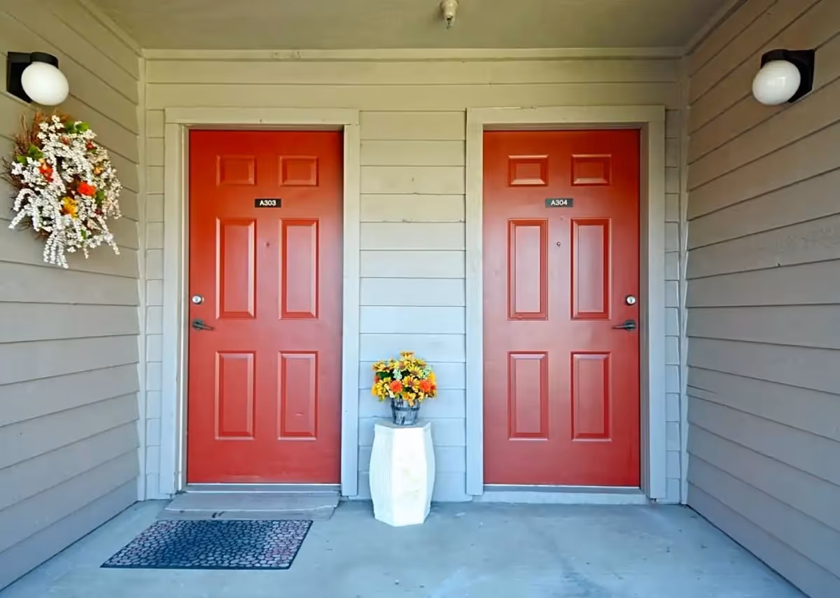 Two red doors labeled A303 and A304 on a beige exterior wall with a flower wreath hanging next to door A303 and a white vase with yellow and orange flowers placed between the doors under two round wall lights.