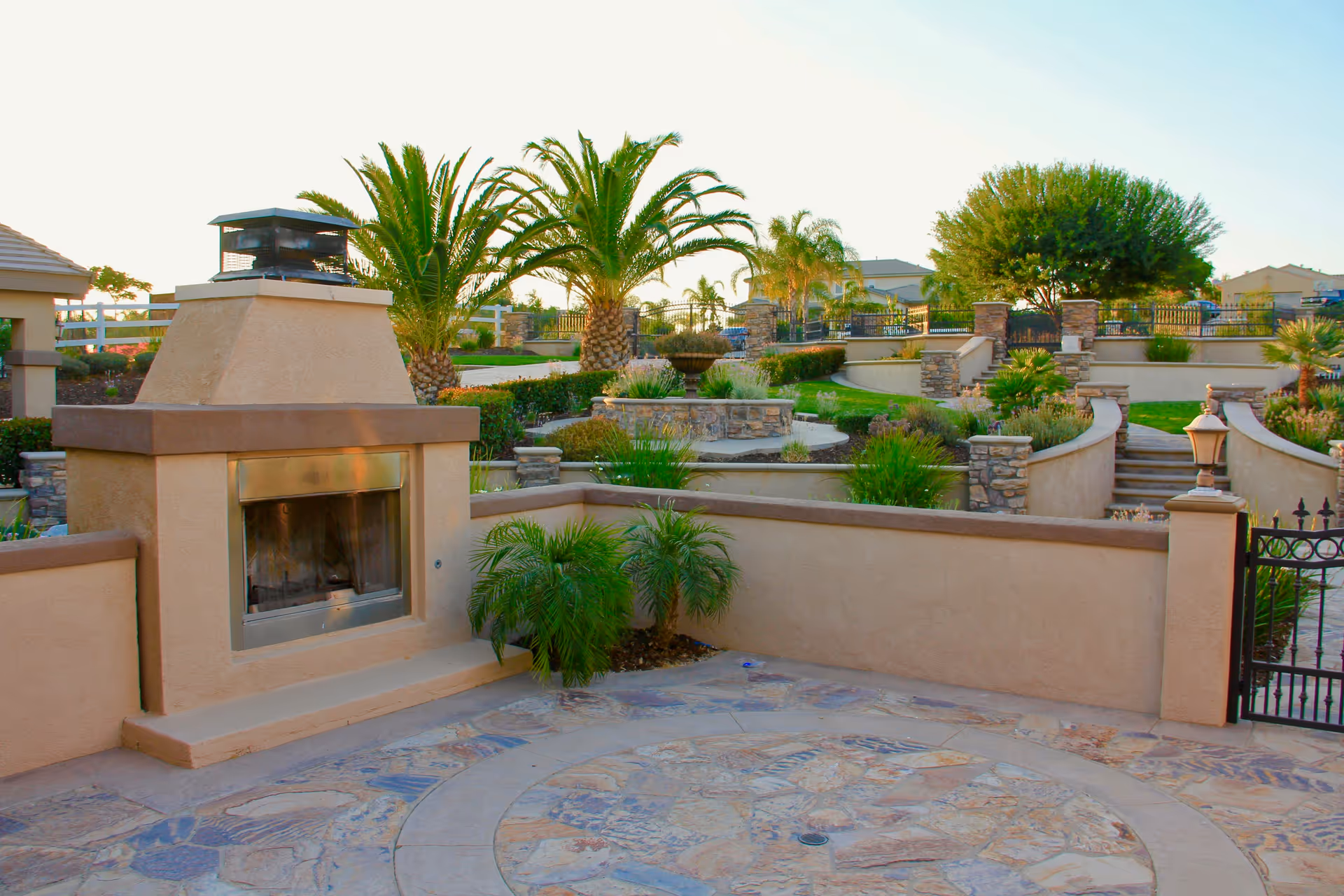 Terraced outdoor courtyard with a built-in fireplace, stone patio, palm trees, and landscaped planters.
