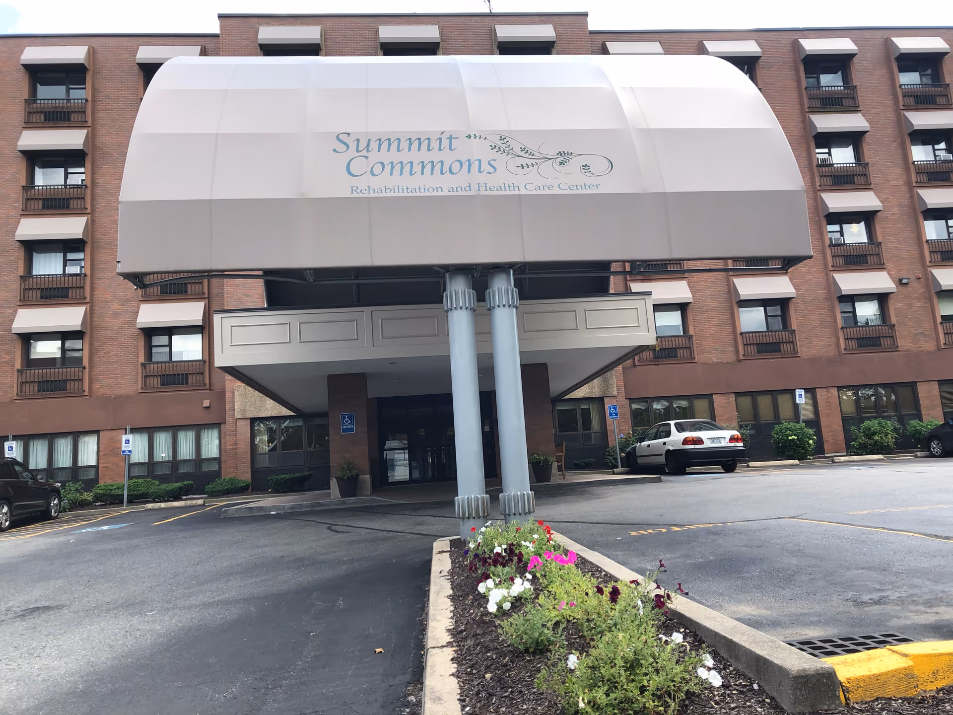 Front exterior view of Summit Commons Rehabilitation and Health Care Center showing a multi-story brick building with a large covered entrance canopy. There are several windows with awnings, a driveway, parking spaces including handicapped spots, and a flower bed with colorful flowers in front.
