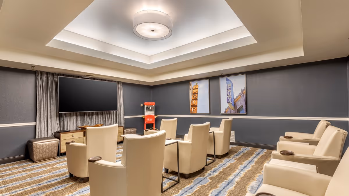 A cozy lounge room with cream-colored armchairs arranged in two rows facing a large flat-screen TV mounted on a dark gray wall. The room features a patterned carpet with blue and beige stripes, a popcorn machine in the corner, and two framed pictures on the wall. The ceiling has a recessed light fixture with soft lighting.