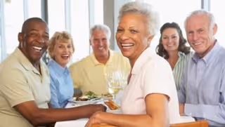 Six smiling older adults seated around a dining table with plates and wine glasses.