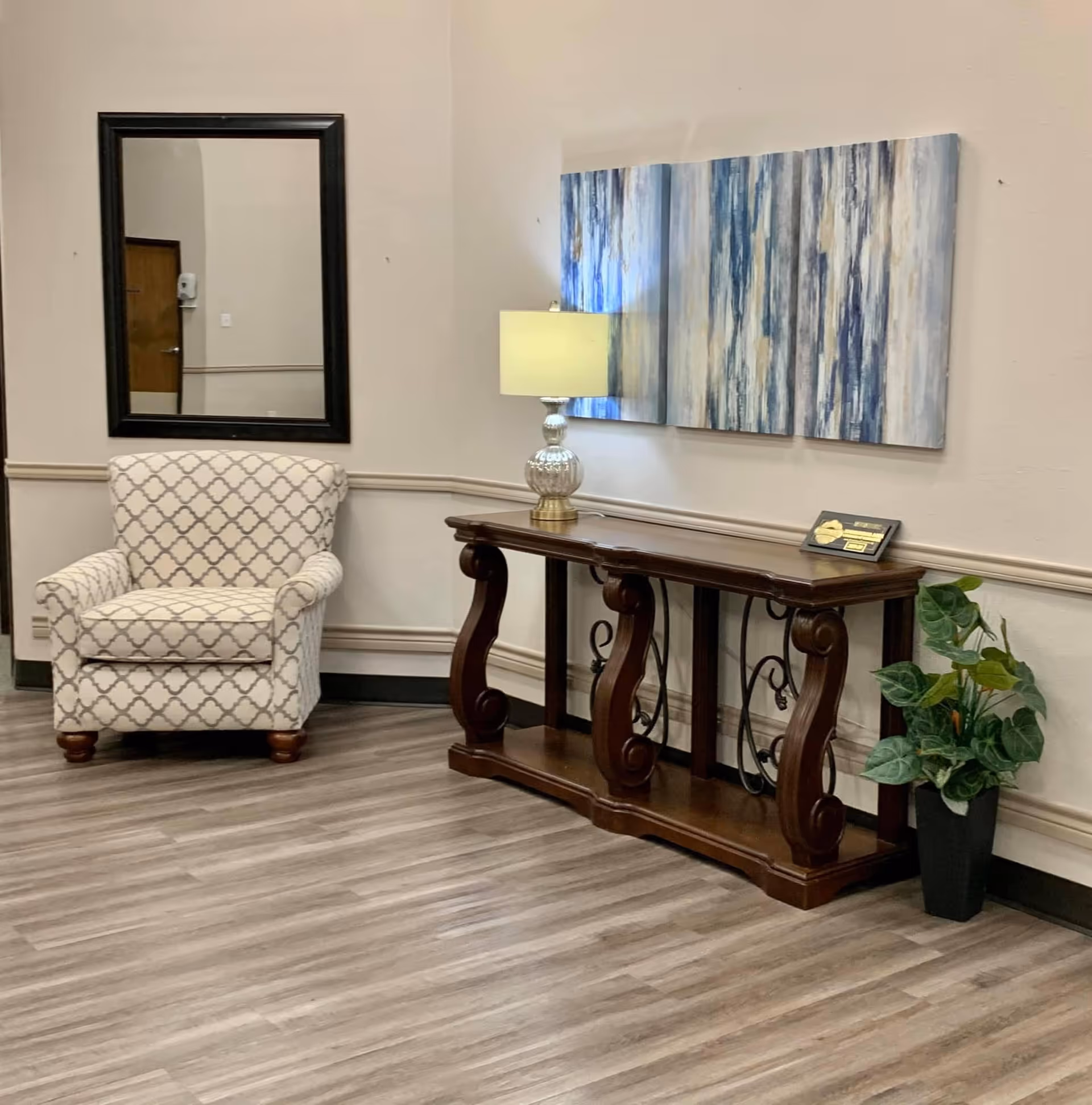 A cozy corner of an interior room featuring a patterned armchair, a wooden console table with ornate legs, a table lamp with a glass base, a three-panel abstract blue and white painting on the wall, a large rectangular mirror, and a potted green plant on the floor.