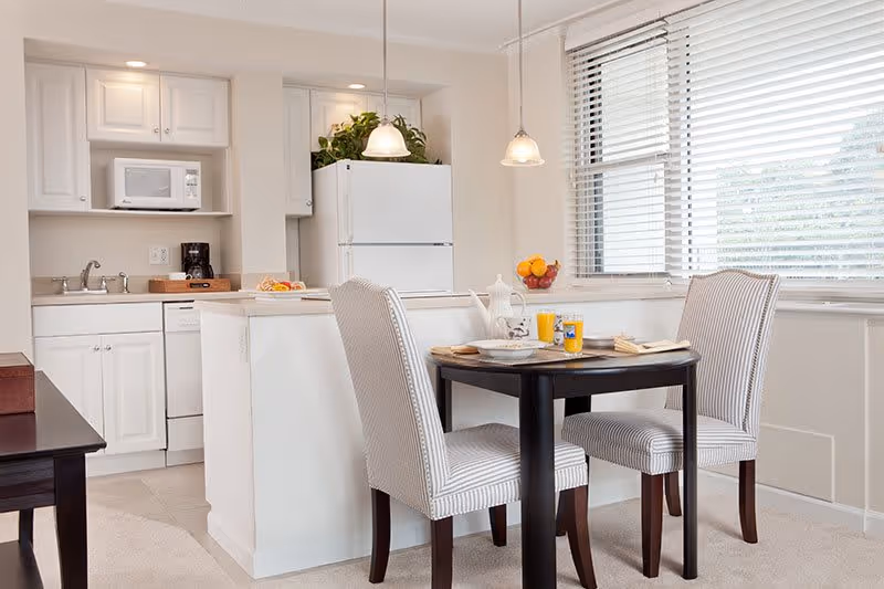 A bright kitchen and dining area with white cabinetry, a white refrigerator, microwave, and dishwasher. There is a small round dining table with two striped upholstered chairs, set with plates, glasses of orange juice, and a teapot. Large windows with white blinds allow natural light to fill the space.
