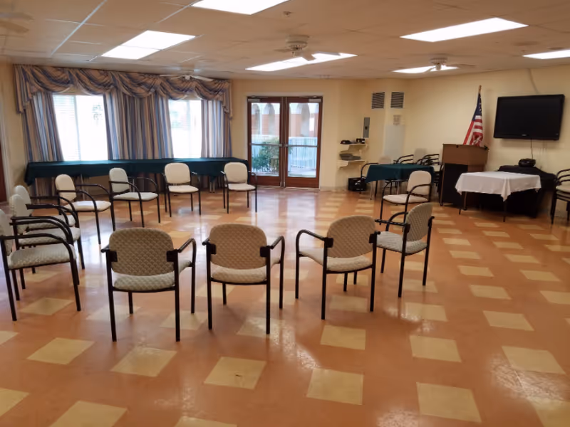 A spacious room with a checkered floor pattern, arranged with chairs in a semi-circle facing a podium and a wall-mounted television. There are tables covered with green and white cloths along the walls, an American flag next to the podium, and large windows with curtains letting in natural light.