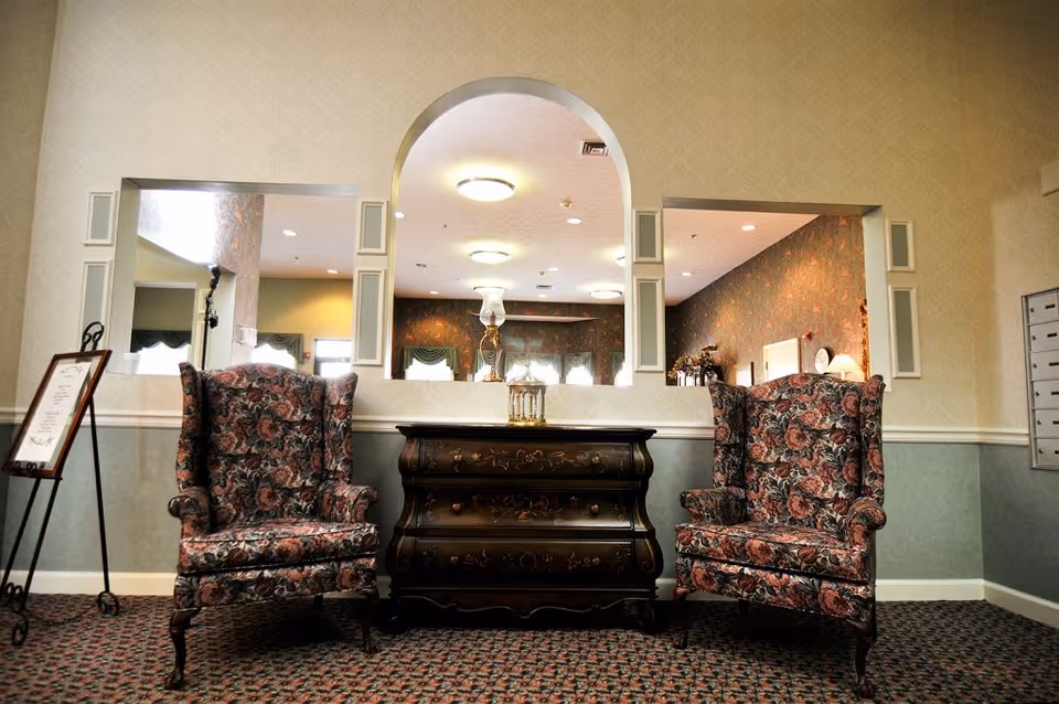 Two floral wingback chairs flank an ornate wooden chest beneath an arched interior opening in a facility sitting area.