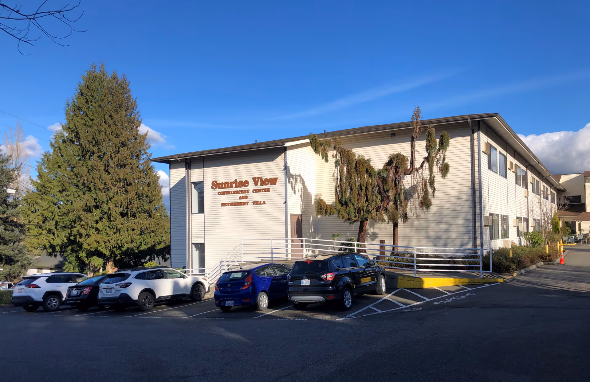 Exterior view of a two-story Sunrise View assisted living building with parked cars and trees under a clear blue sky.