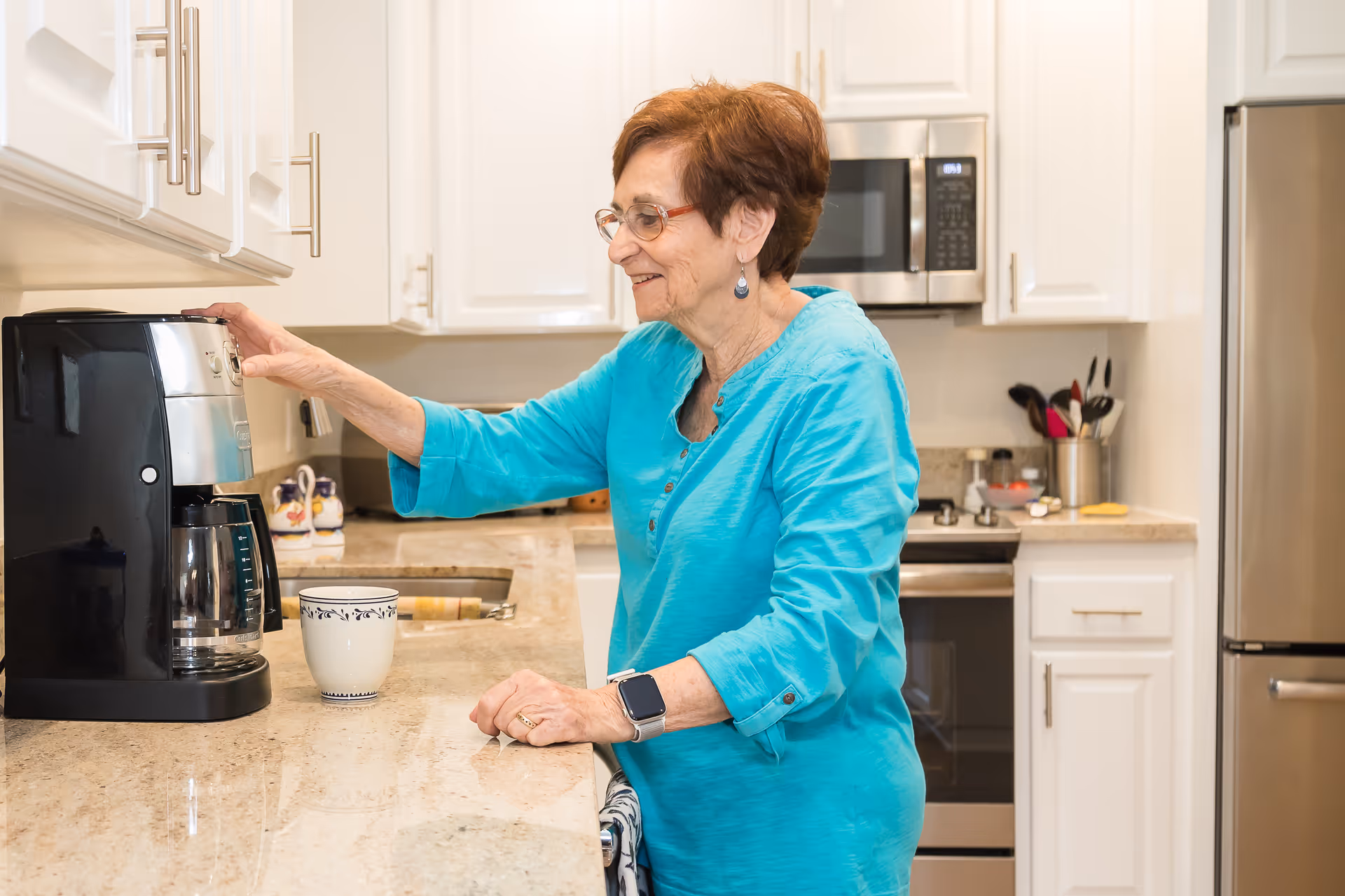 An elderly woman wearing glasses and a turquoise shirt is making coffee using a black coffee maker in a modern kitchen with white cabinets and granite countertops. A white mug with a blue pattern is placed on the counter next to the coffee maker.