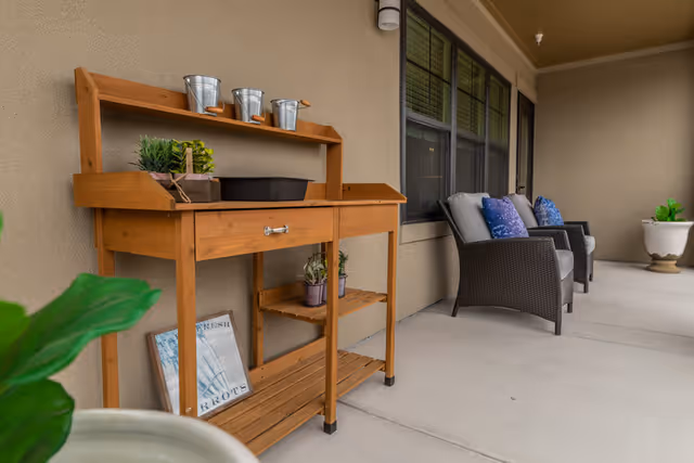 Covered outdoor patio area with a wooden potting bench holding small plants and metal buckets, two cushioned wicker chairs with blue pillows, and large potted plants along the wall of a building with windows.