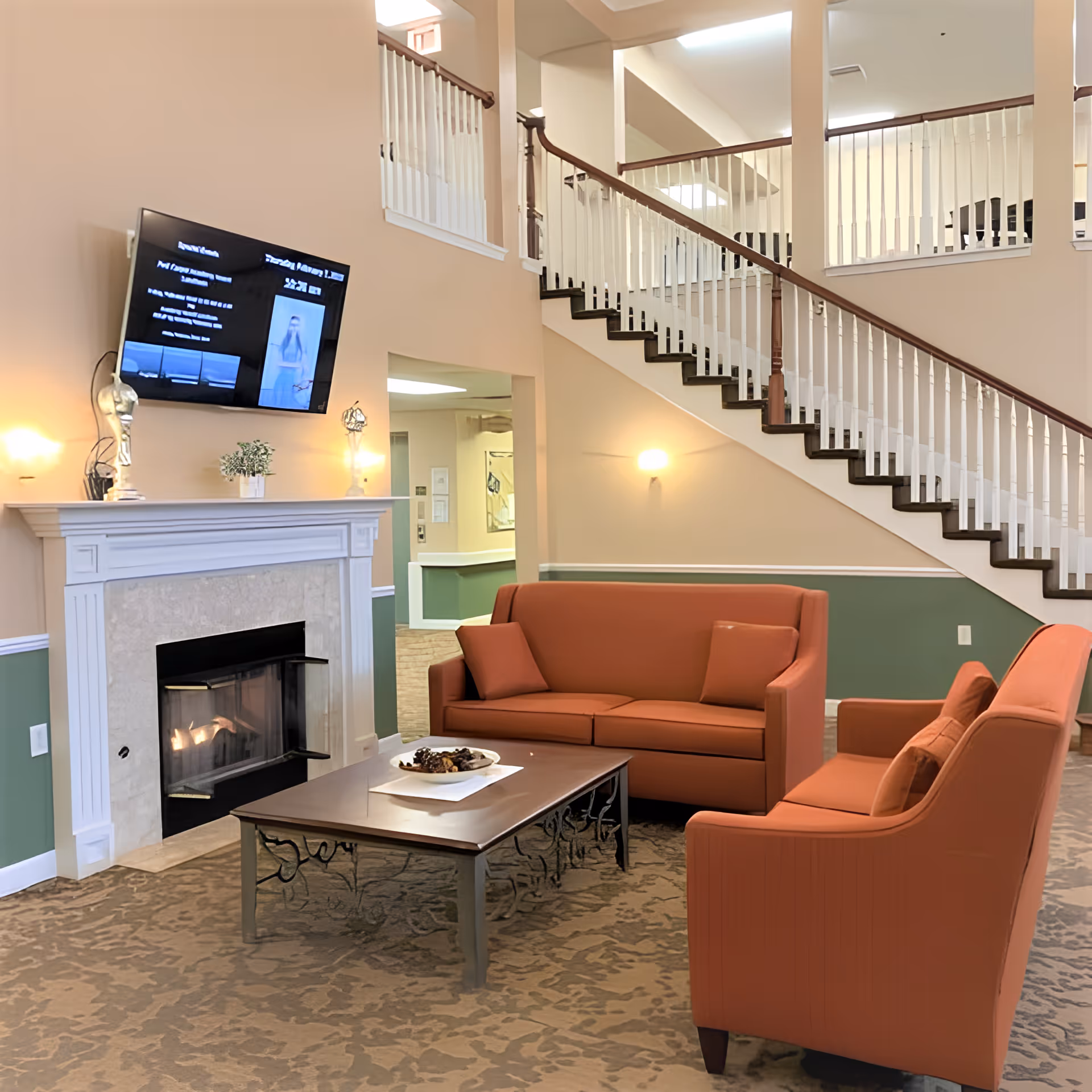 A cozy living room area with two orange sofas facing a wooden coffee table. Behind the sofas is a white fireplace with a fire burning and a flat-screen TV mounted above it. The room features a staircase with white railings and a beige and green wall color scheme.
