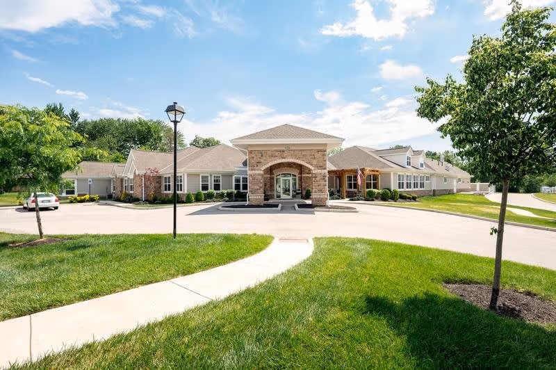 Front exterior view of a single-story senior living facility building with a covered entrance, surrounded by green grass, trees, and a clear blue sky with some clouds.