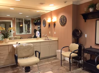 Interior view of a salon or grooming area in an assisted living facility, featuring a salon chair in front of a large mirror and countertop with a sink. There are two wall clocks, wall-mounted light fixtures, a hair dryer station, and a small waiting area with two chairs. The room has warm lighting and wood-patterned flooring.