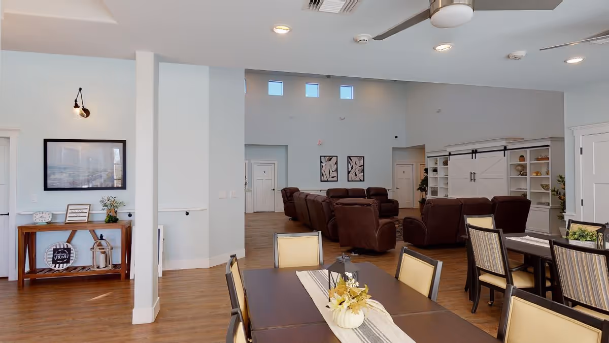 A spacious and well-lit common area in an assisted living facility featuring a dining table with chairs in the foreground and several brown recliner chairs arranged in a seating area in the background. The room has light-colored walls, wooden flooring, decorative wall art, and built-in shelving with sliding barn-style doors.