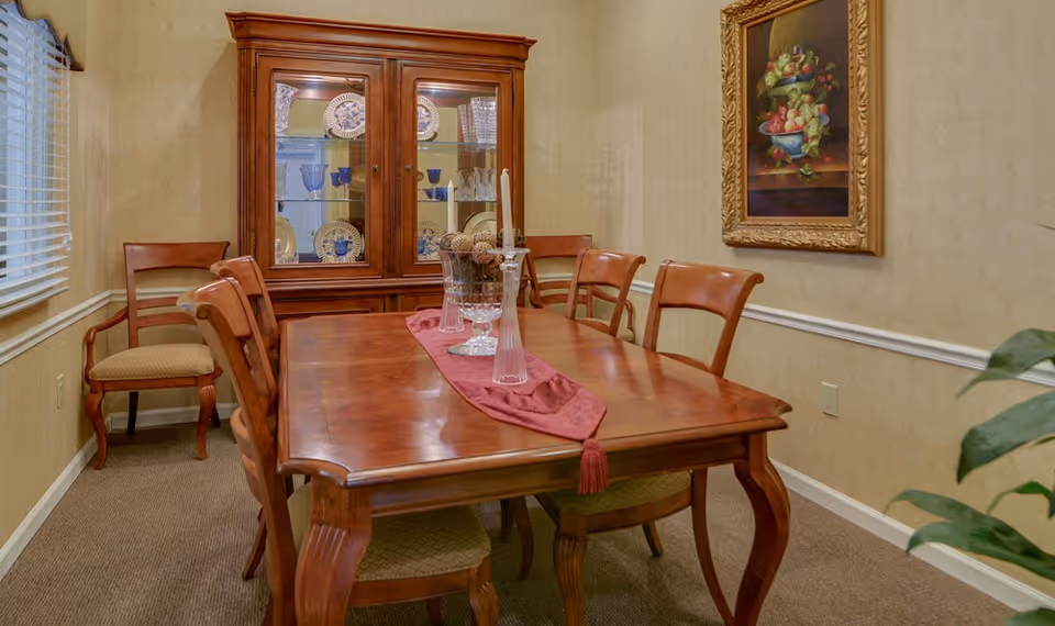 A traditional dining room with a wooden dining table and six matching chairs. The table is decorated with a red runner and glass candle holders. A wooden china cabinet with glass doors displays plates and glassware. A framed painting of fruit hangs on the wall, and a window with blinds is on the left side.
