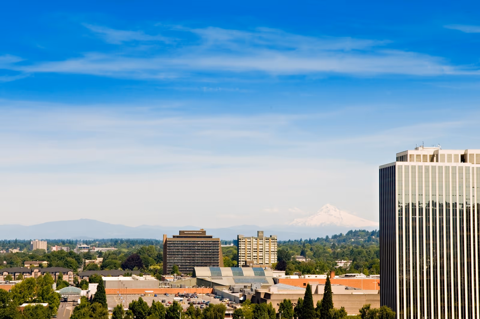 Cityscape view featuring multiple buildings, a parking lot, and a distant snow-capped mountain under a blue sky with scattered clouds.